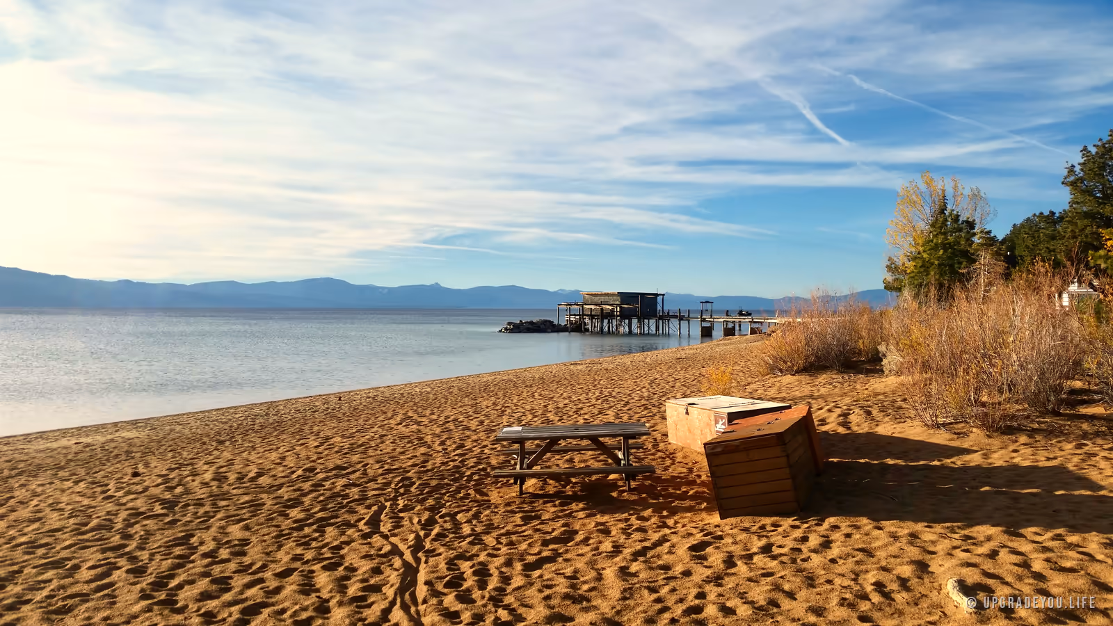 A calm blue alpine lake meeting a sandy shore with distant mountains under a wide sky