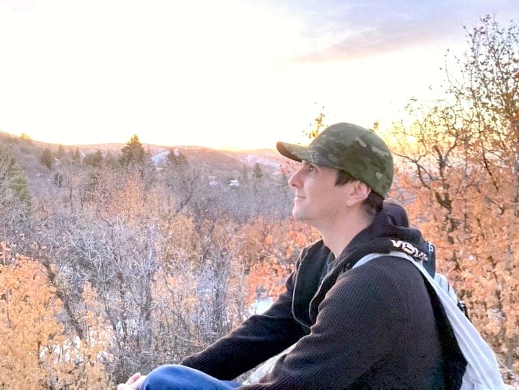 Jess seduto su una collina al tramonto, con lo sguardo su querce autunnali e neve in lontananza.