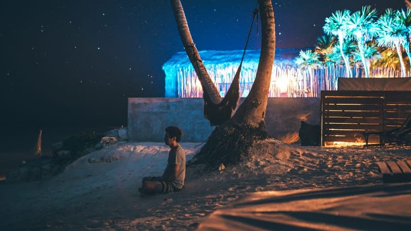 Jess seduto a gambe incrociate sulla sabbia sotto una palma di notte, stelle sopra una capanna illuminata sulla spiaggia.